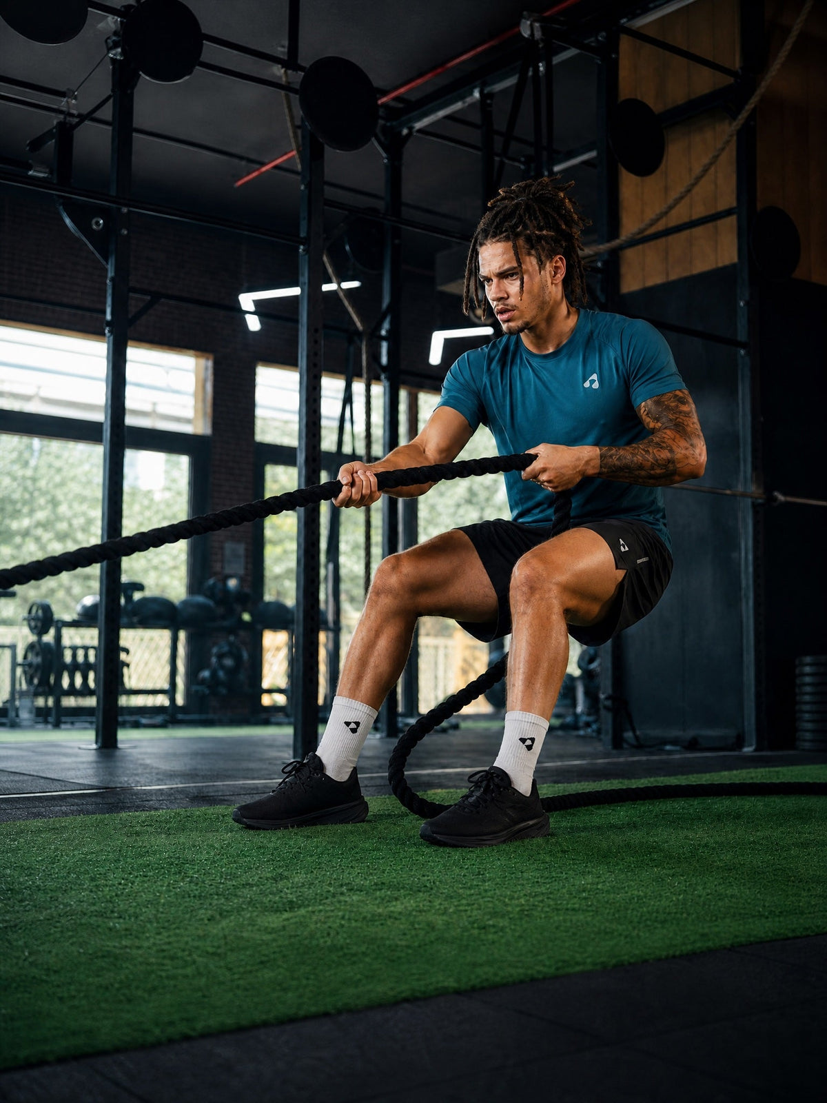 Man wearing a blue Boost Sportswear performance T-shirt and black training shorts while exercising with battle ropes in the gym.